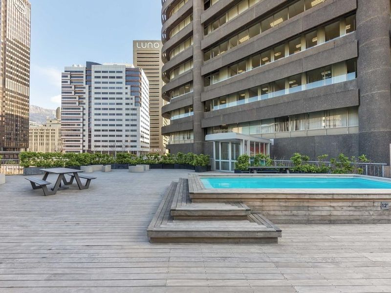A rooftop terrace featuring a raised timber deck and a built-in swimming pool, set against a backdrop of city high-rise buildings.