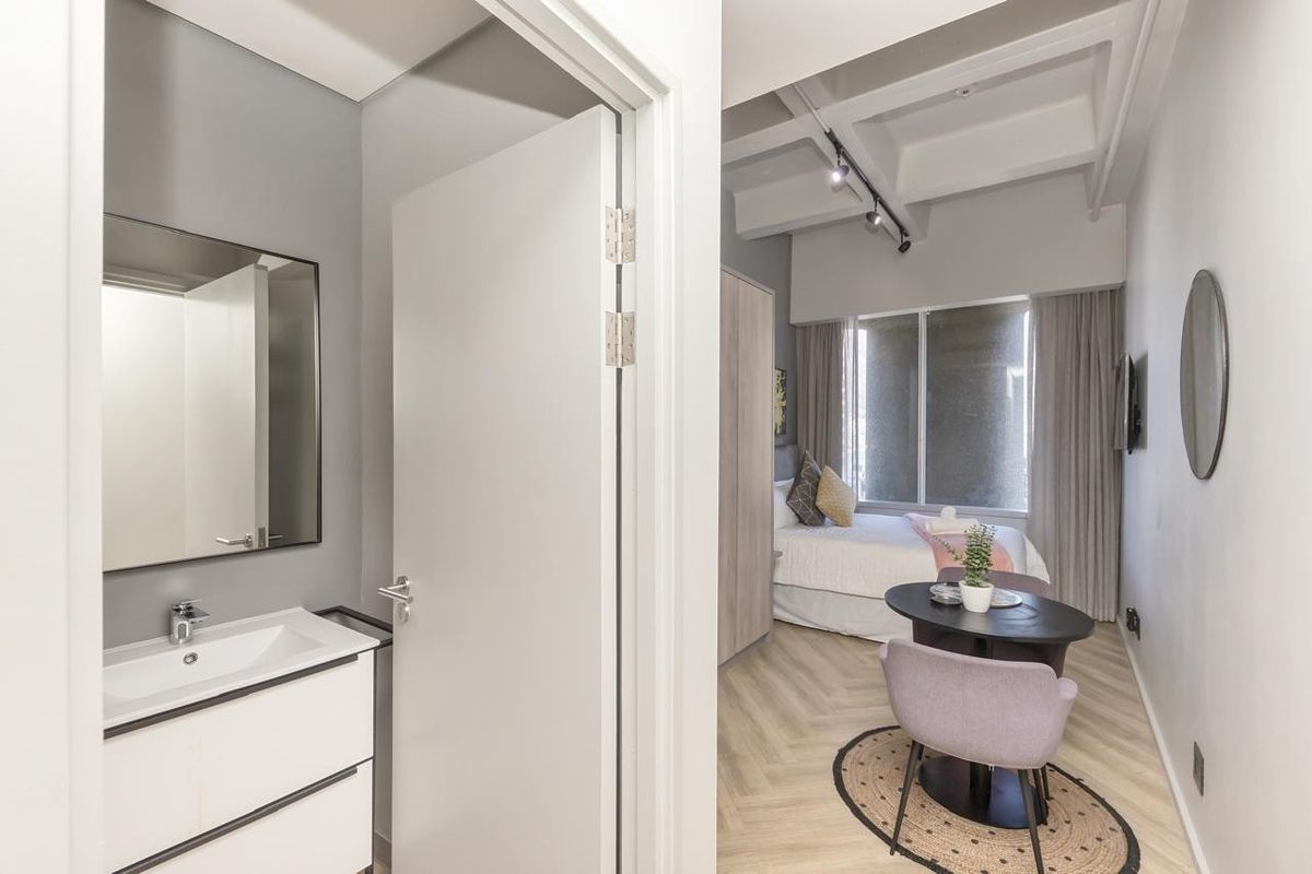 A modern bedroom featuring herringbone flooring, track lighting, and an en-suite vanity area with a contemporary basin and mirror.