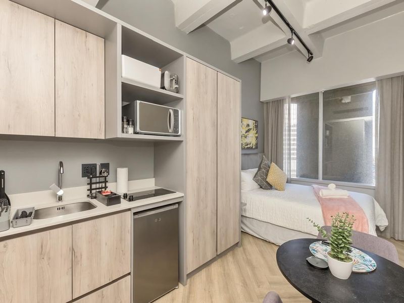 Modern open-plan studio apartment featuring a compact kitchenette with light wood cabinetry, integrated appliances, and a combined living and sleeping area with neutral finishes.