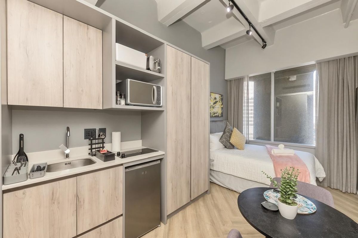 Modern open-plan studio apartment featuring a compact kitchenette with light wood cabinetry, integrated appliances, and a combined living and sleeping area with neutral finishes.