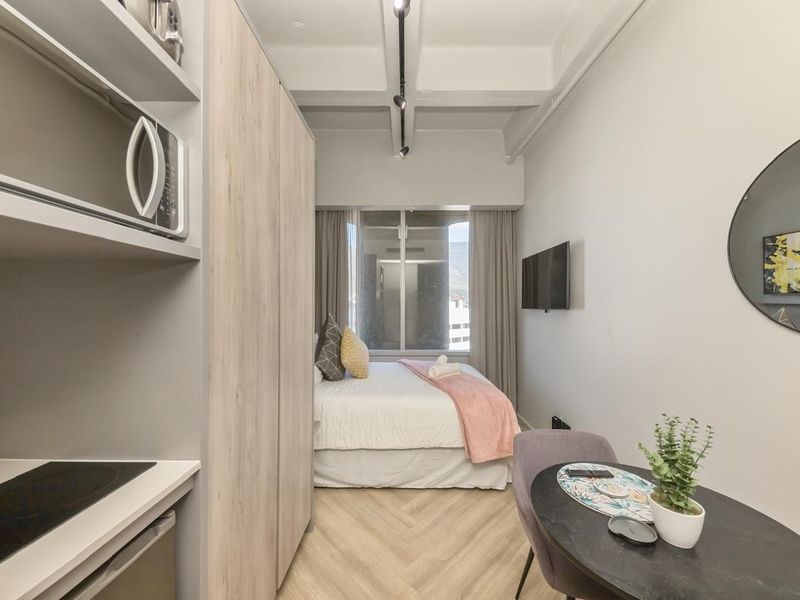 A modern open-plan studio apartment featuring herringbone flooring, exposed ceiling beams, built-in kitchen cabinetry with integrated appliances, and a compact sleeping area.