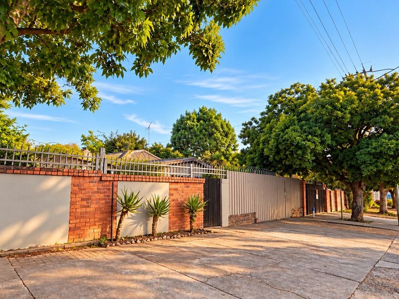 Front exterior view of the property featuring a brick boundary wall, metal security gate, and concrete driveway shaded by mature trees.