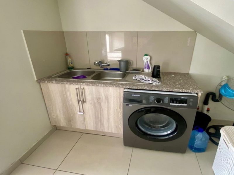 A compact laundry area featuring a double sink, laminate countertop, built-in cabinetry, and space for a washing machine under a sloped ceiling.