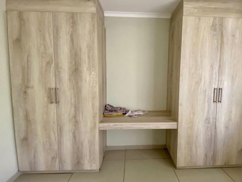 A bedroom featuring light wood-grain built-in cupboards with a central vanity desk and neutral tiled flooring.
