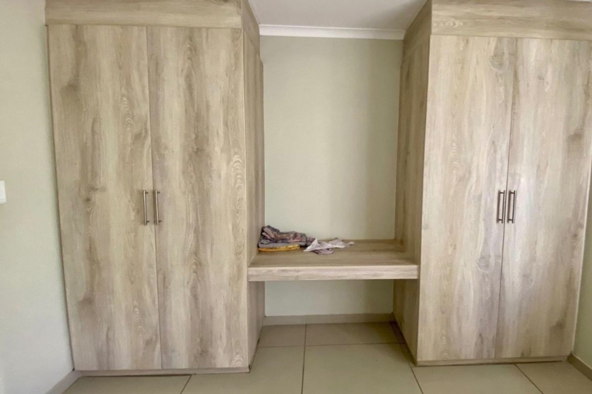 A bedroom featuring light wood-grain built-in cupboards with a central vanity desk and neutral tiled flooring.