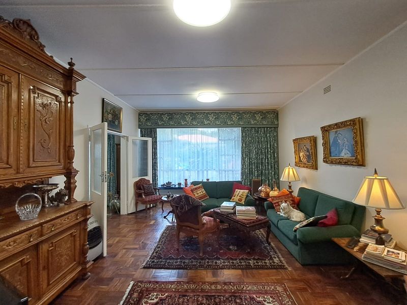A spacious living room featuring classic parquet flooring, large windows with decorative pelmets, and traditional ceiling lighting.