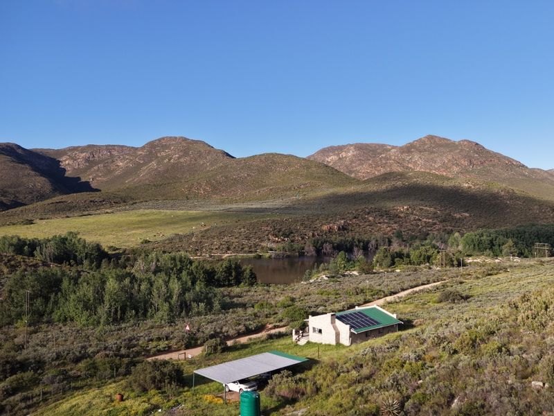 Aerial view of a rural farm property featuring a main dwelling with solar panels, a detached carport, and a water tank set against mountain scenery.