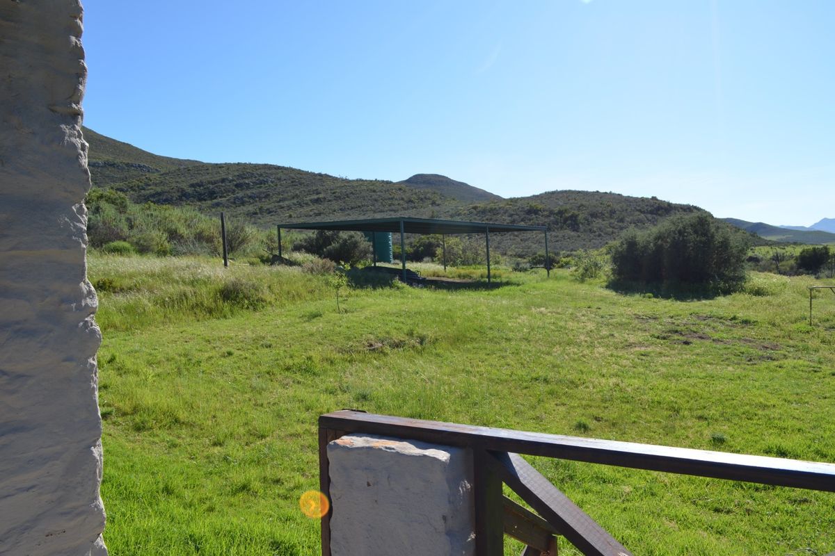 A view of the rear farm grounds featuring a metal carport structure, stone pillar, and timber railing set against a natural landscape.