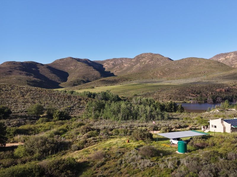 Aerial view of a rural farm property featuring a dwelling with solar panels, a carport, and a water storage tank set against mountain scenery.