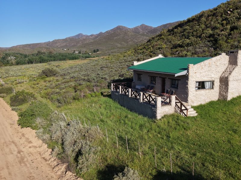 Aerial view of a rustic stone cottage with a green metal roof, featuring a raised patio and scenic mountain landscape.