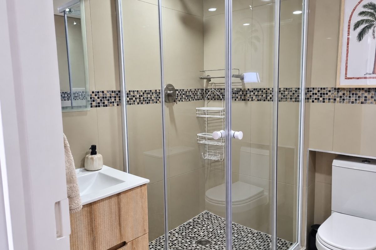 A modern bathroom featuring a glass-enclosed shower, a wood-finish vanity with integrated basin, and a toilet, accented with decorative mosaic tile detailing.