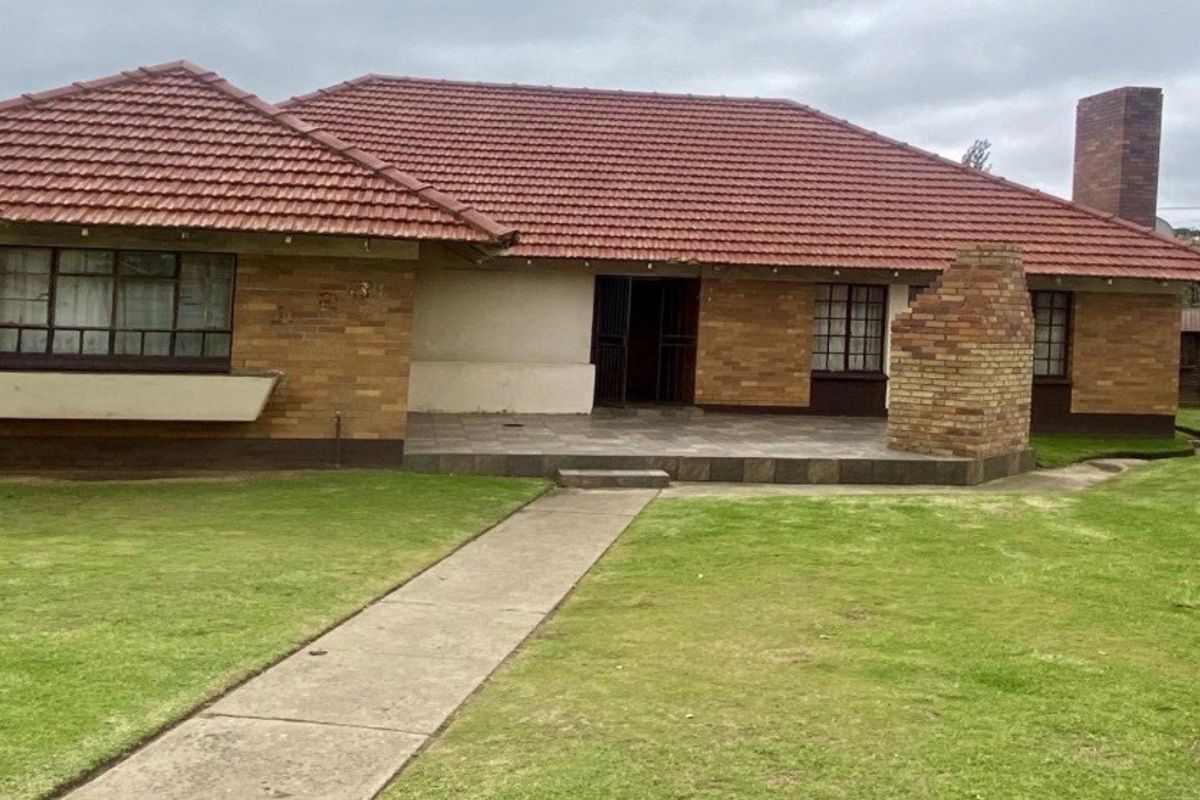 Rear view of a single-story brick house featuring a tiled roof, a paved patio area, and a spacious lawn with a concrete walkway.