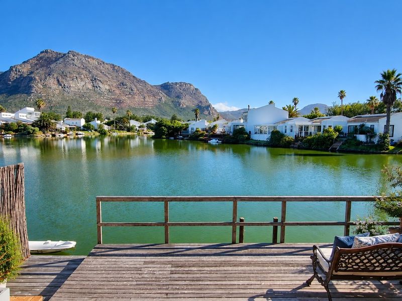 A scenic view from a wooden deck overlooking a tranquil lake with surrounding residential properties and mountain vistas in the background.