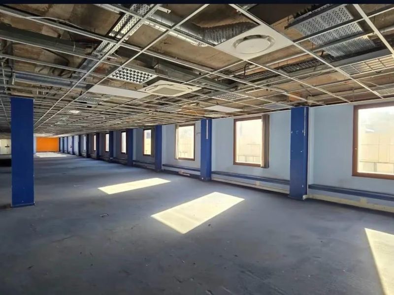 A large, open-plan office space currently undergoing renovation, featuring exposed ceiling infrastructure, structural columns, and natural light from a row of windows.