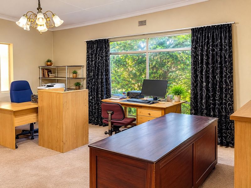 A functional home office space featuring large windows with garden views, neutral wall tones, and carpeted flooring.