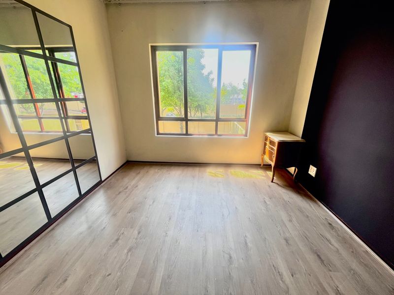 A bright, empty bedroom featuring modern laminate flooring, a large built-in mirror wardrobe, and a window providing natural light.