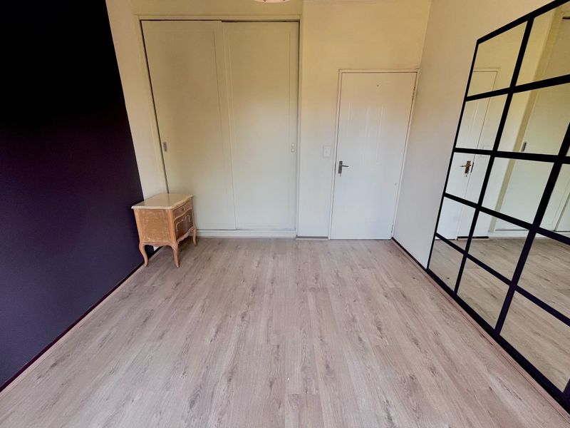 A bright, empty bedroom featuring light-toned laminate flooring, a built-in sliding wardrobe, and a dark accent wall.
