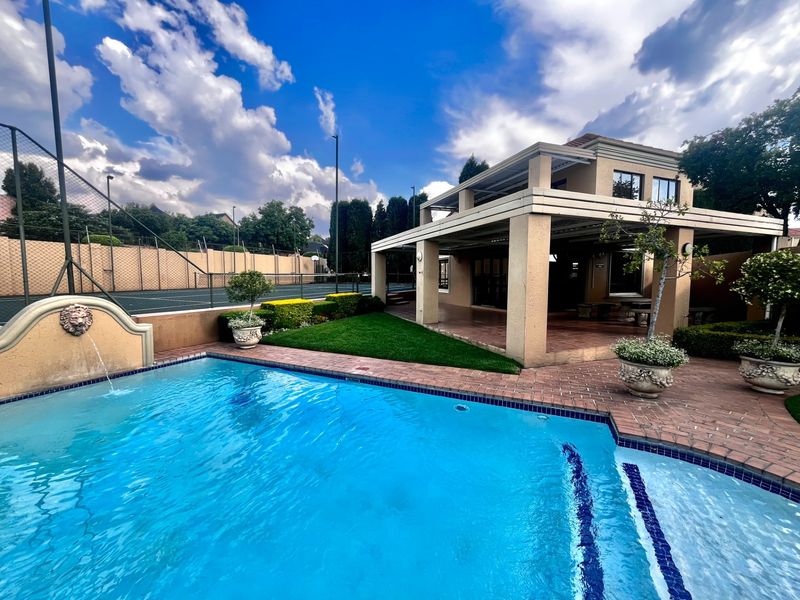 A well-maintained outdoor entertainment area featuring a swimming pool with a water feature, a covered patio, and an adjacent tennis court.