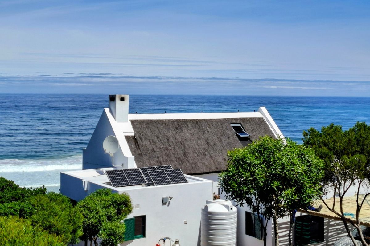 Aerial view of a coastal property featuring a traditional thatch roof, integrated solar panels, and a water storage tank overlooking the ocean.