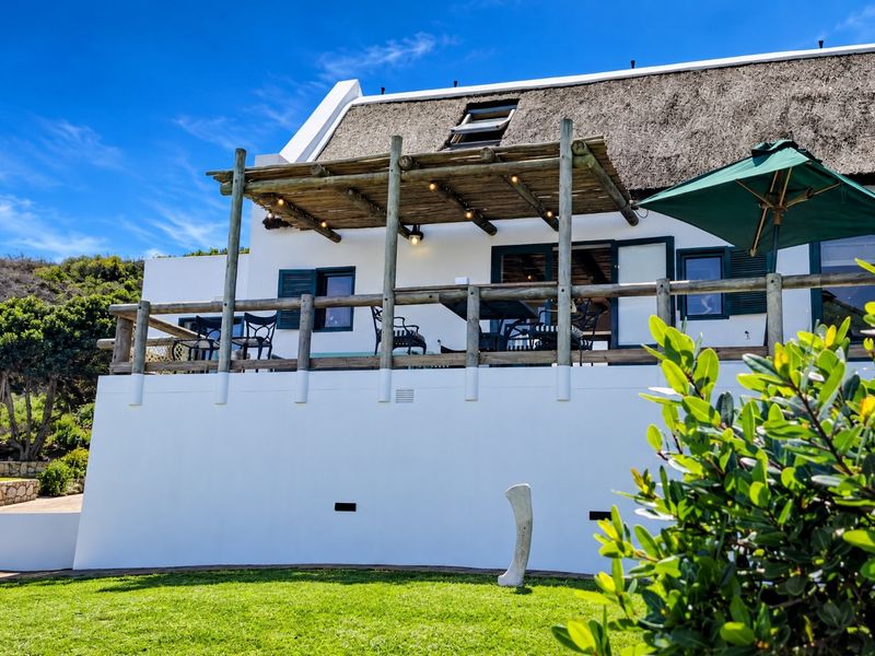 Rear elevation of a charming thatched-roof house featuring a raised timber deck with a pergola, overlooking a well-maintained garden.
