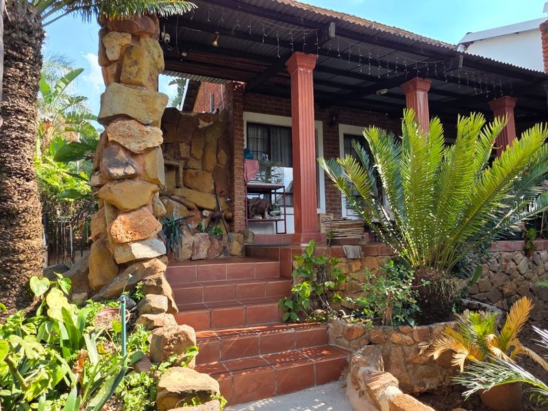 A rustic covered patio area featuring stone-clad pillars, tiled steps, and a corrugated metal roof, set within a landscaped garden environment.