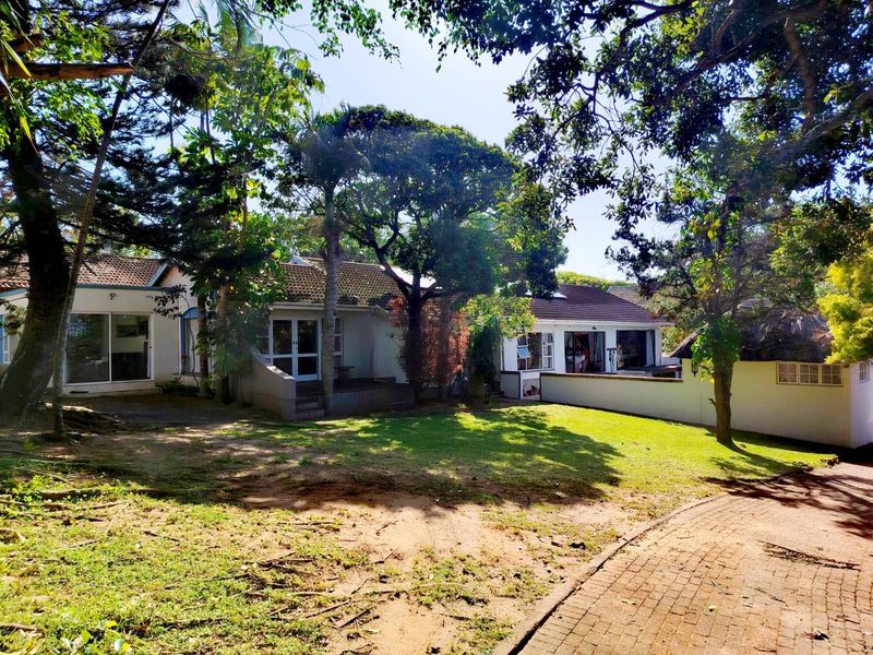 A spacious rear garden view featuring a well-maintained lawn, mature trees, paved driveway, and the rear facade of the house with sliding door access.