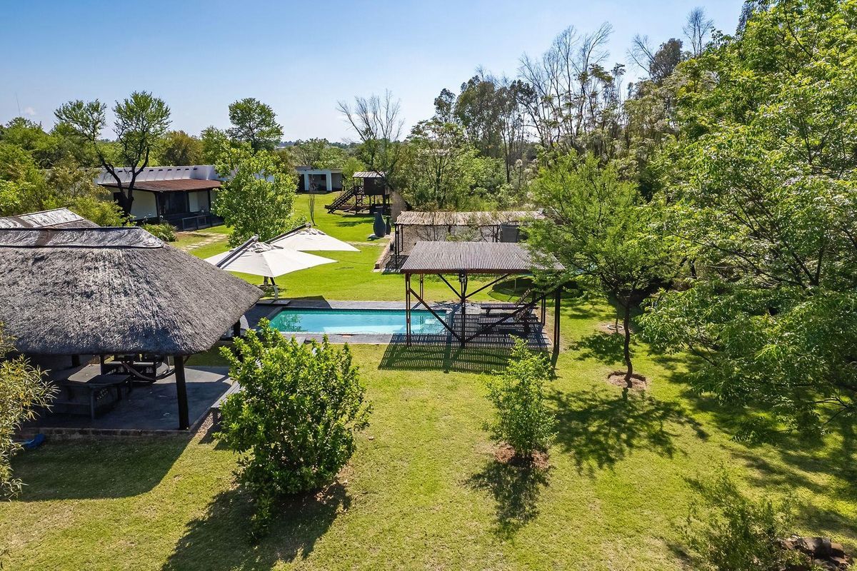 Aerial view of a spacious farm property featuring a swimming pool, a traditional thatched lapa, and a timber pergola set within a lush garden.