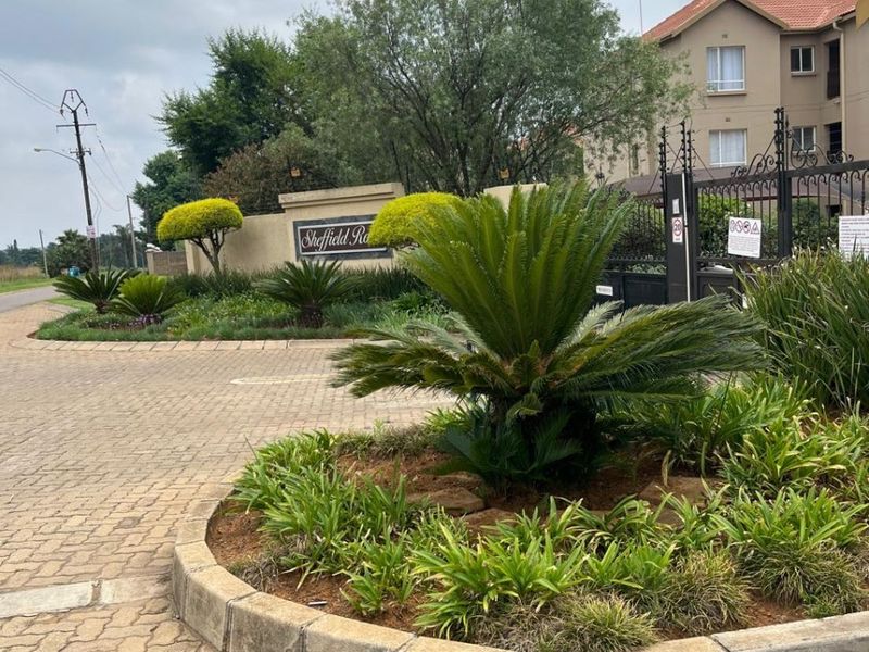 Entrance view of the townhouse complex featuring a paved driveway, landscaped garden beds, and secure gated access.