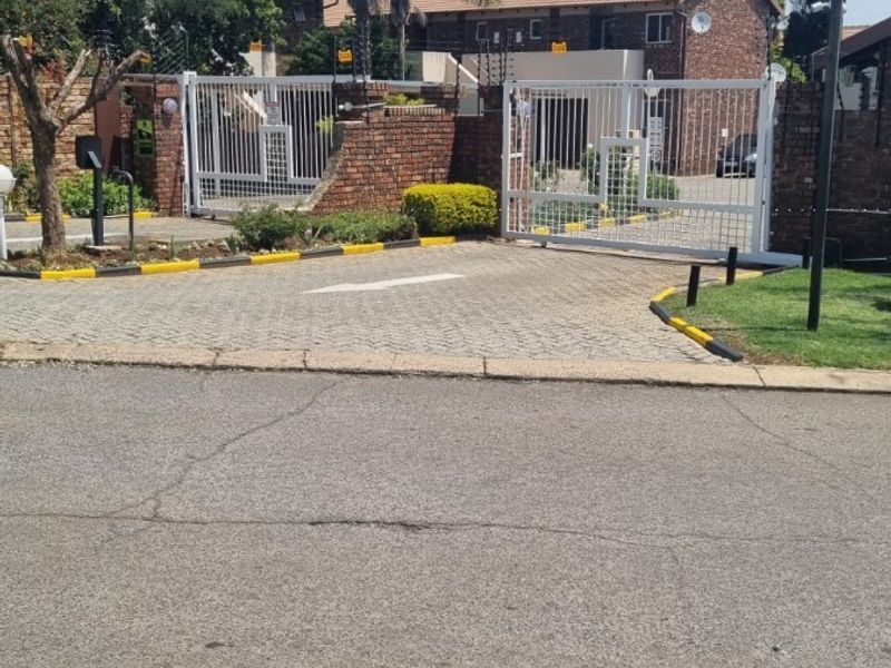 Front view of the apartment complex entrance featuring a paved driveway, secure metal gates, brick boundary walls, and electric fencing.