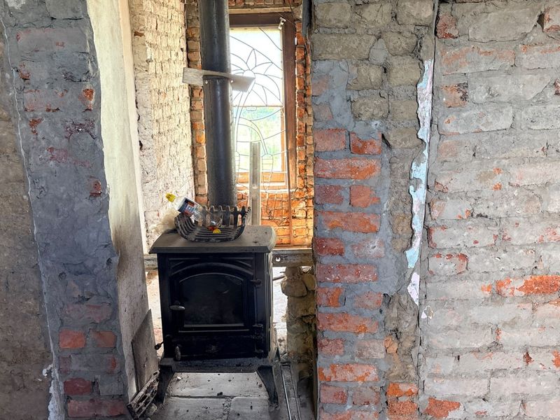 An interior space under renovation featuring exposed brick walls, a cast iron wood burning stove, and a decorative leaded glass window.