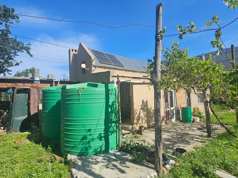 Rear exterior view of the property featuring solar panels, water storage tanks, and a concrete patio area with surrounding garden space.