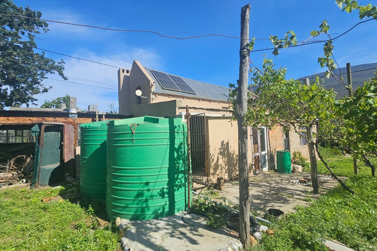Rear exterior view of the property featuring solar panels, water storage tanks, and a concrete patio area with surrounding garden space.