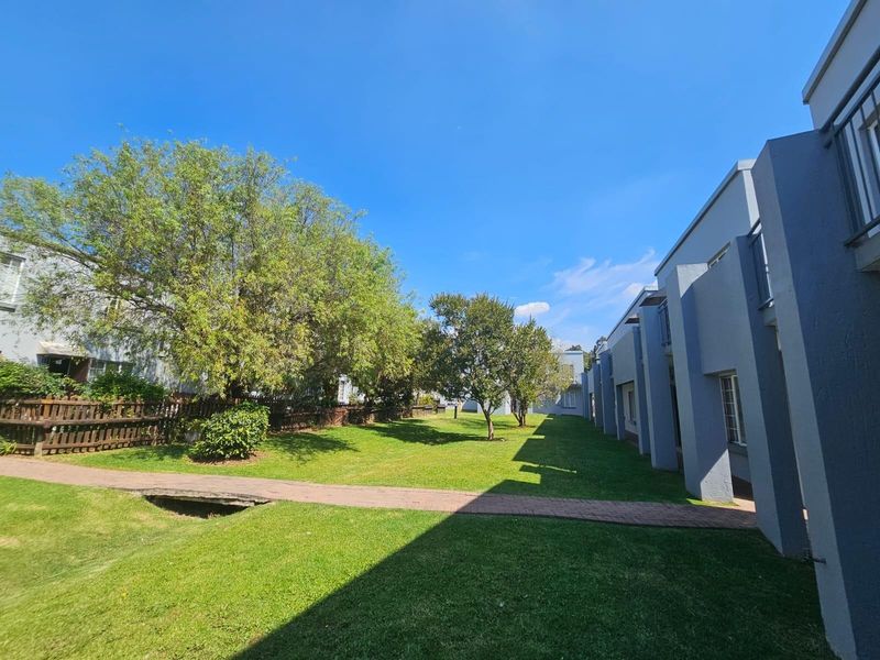 A well-maintained communal garden area featuring a paved walkway, mature trees, and lawn space situated between townhouse units.