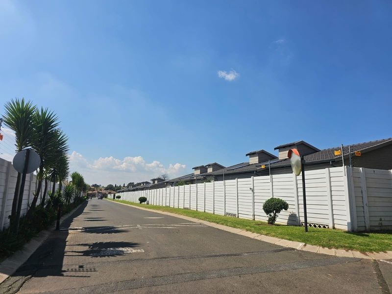 A view of the residential street showing the townhouse complex boundary wall, security fencing, and paved road access.