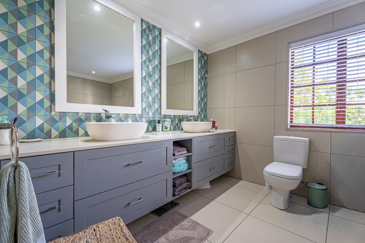 Modern bathroom featuring a double vanity with vessel sinks, patterned feature wall tiling, ample built-in cabinetry, and a private toilet area.