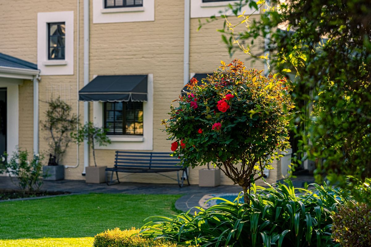 A well-maintained rear exterior view featuring a brick facade, window awnings, and a landscaped garden with a paved patio area.