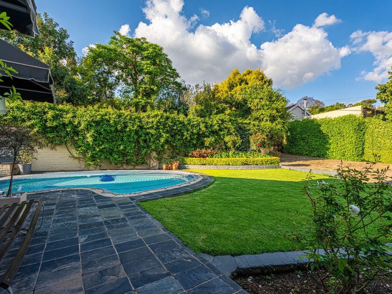 A well-maintained outdoor pool area featuring a stone-paved patio, lush green lawn, and established garden hedges providing privacy.