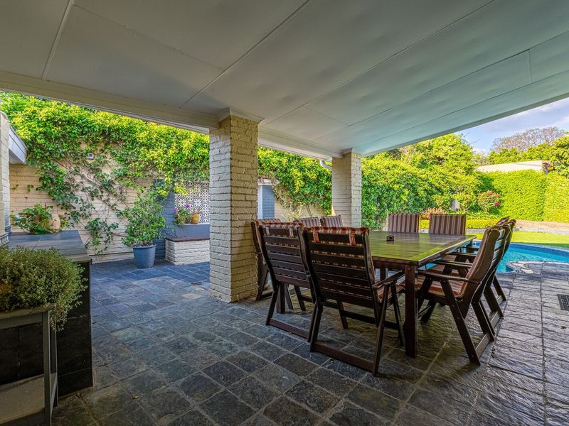 A spacious covered patio featuring stone tile flooring, brick pillars, and a built-in outdoor counter, overlooking a private swimming pool and garden area.
