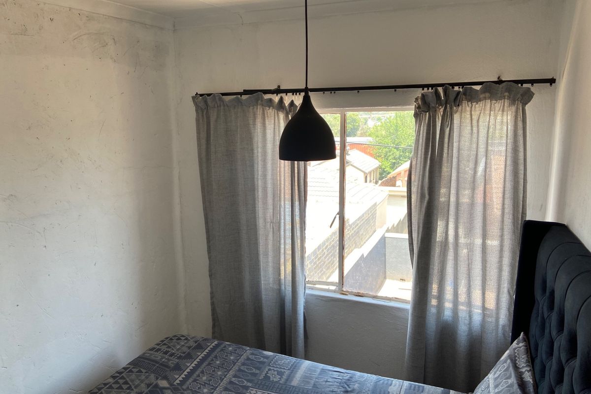 A bedroom featuring a window with curtains and a pendant light, showing signs of wall surface wear and plaster imperfections.