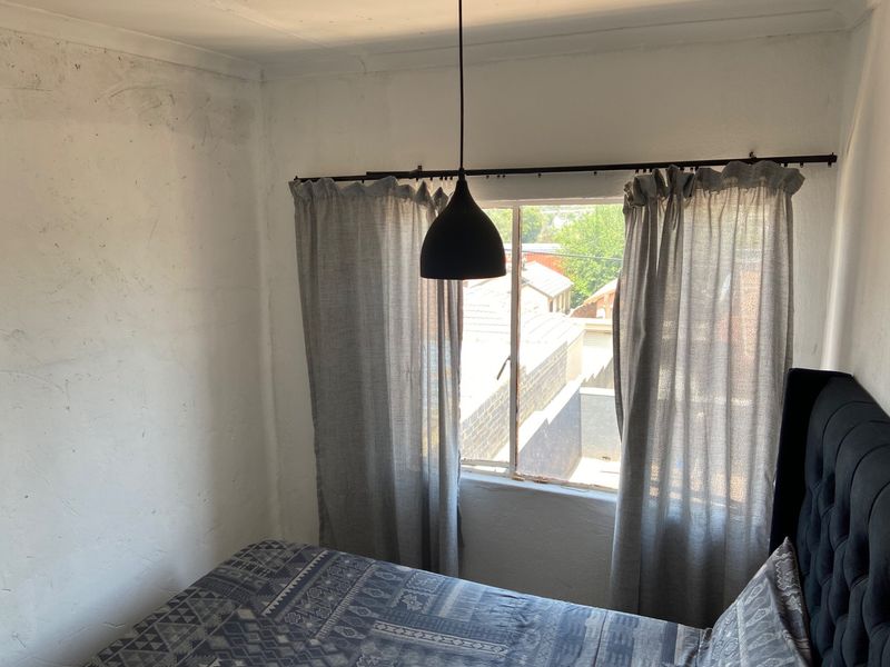 A bedroom featuring a window with curtains and a pendant light, showing signs of wall surface wear and plaster imperfections.