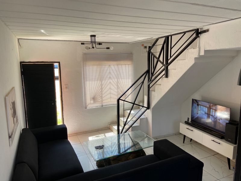 A living room featuring tiled flooring, a staircase with a metal balustrade, and natural light through venetian blinds.