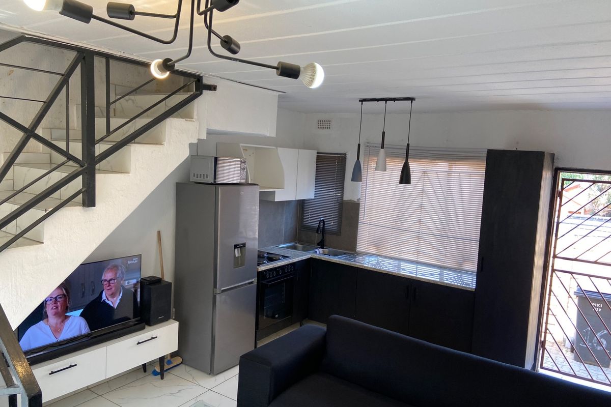 Open-plan living area featuring a compact kitchen with dark cabinetry, tiled flooring, and an integrated staircase leading to the upper level.