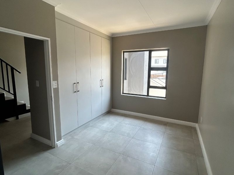 A bright, empty bedroom featuring neutral tiled flooring, built-in cupboards, and a modern aluminium window frame, offering a clean and versatile living space.