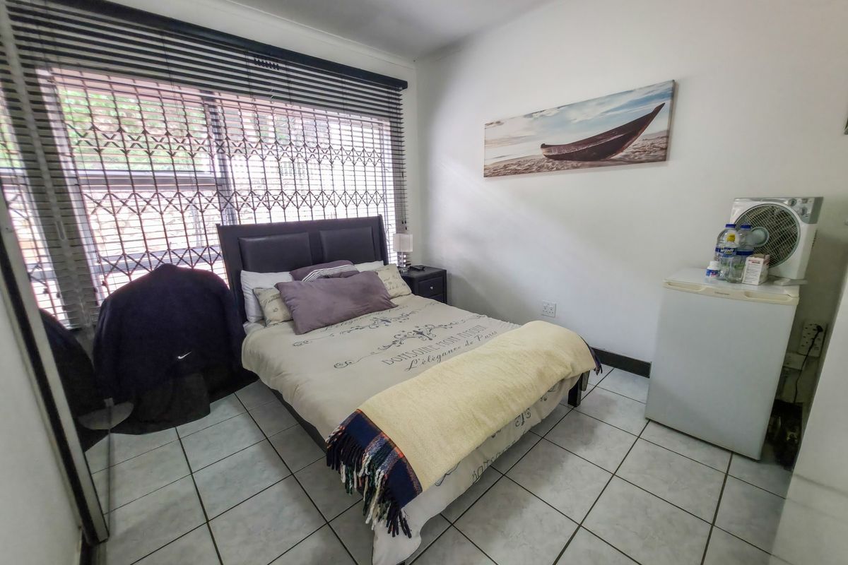 A functional bedroom featuring tiled flooring, a large window with security gate and venetian blinds, providing a clean and practical living space.