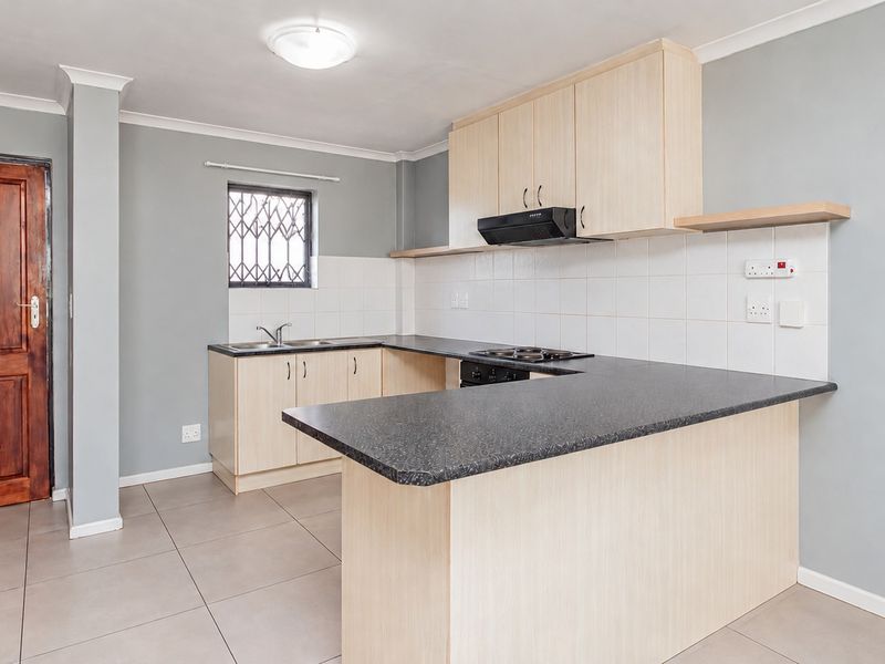 A clean and functional kitchen featuring light wood cabinetry, dark laminate countertops, a built-in hob with extractor fan, and neutral tiled flooring.