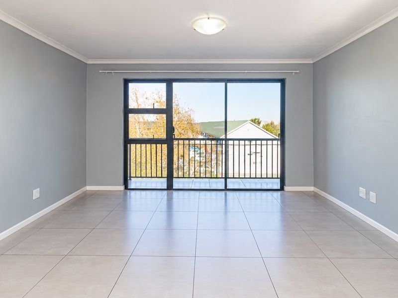 A bright, empty living room featuring neutral tiled flooring, grey walls, and a large sliding glass door leading to a balcony.