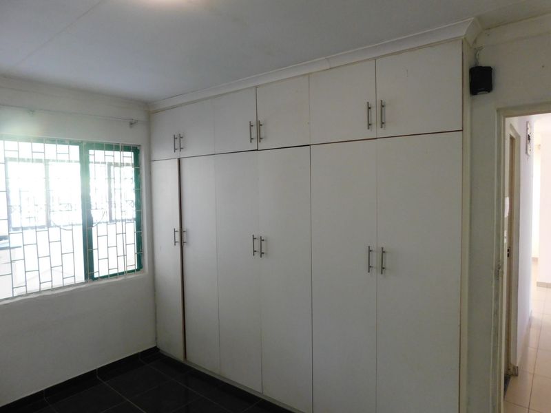 A clean, empty bedroom featuring extensive floor-to-ceiling built-in cupboards, tiled flooring, and a window fitted with security bars.