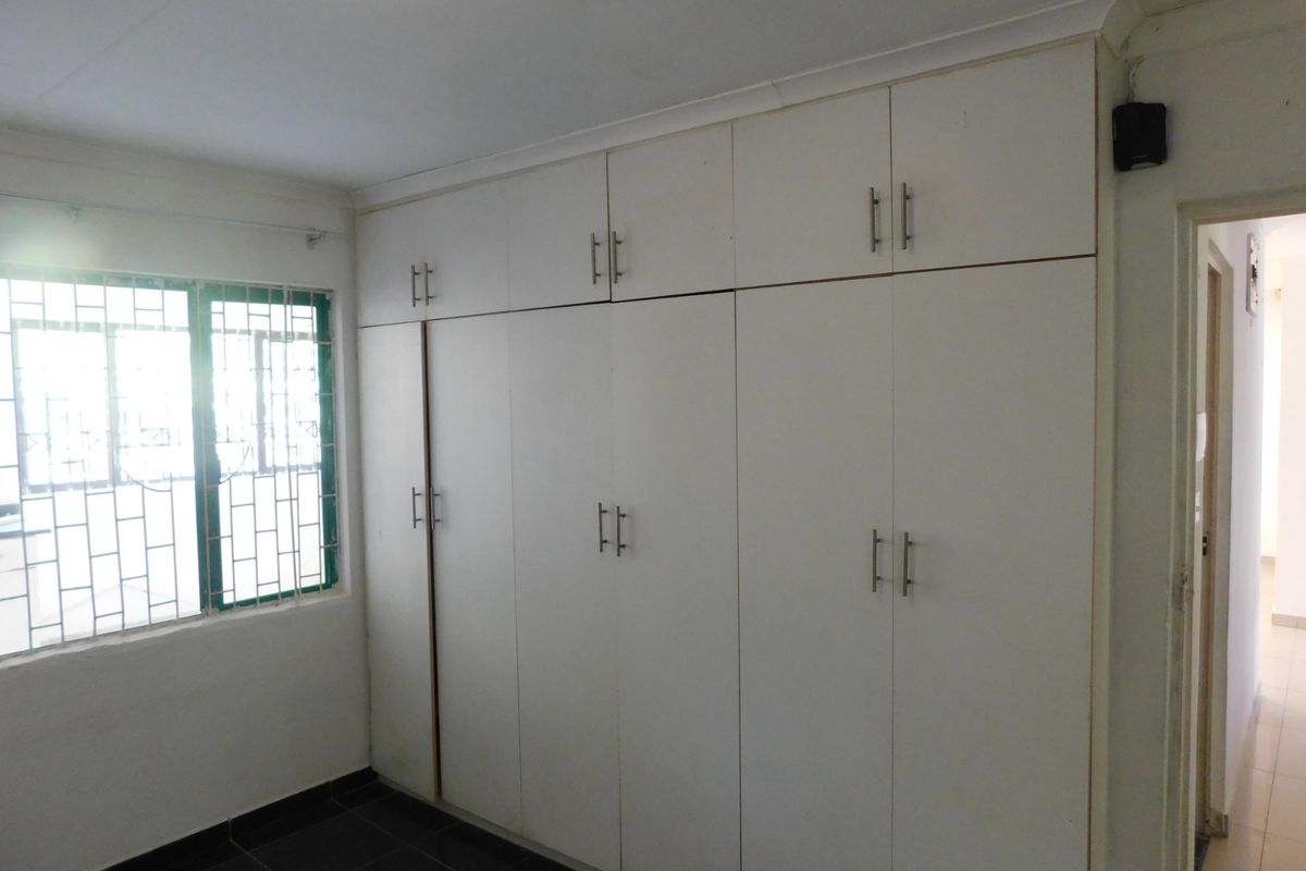 A clean, empty bedroom featuring extensive floor-to-ceiling built-in cupboards, tiled flooring, and a window fitted with security bars.