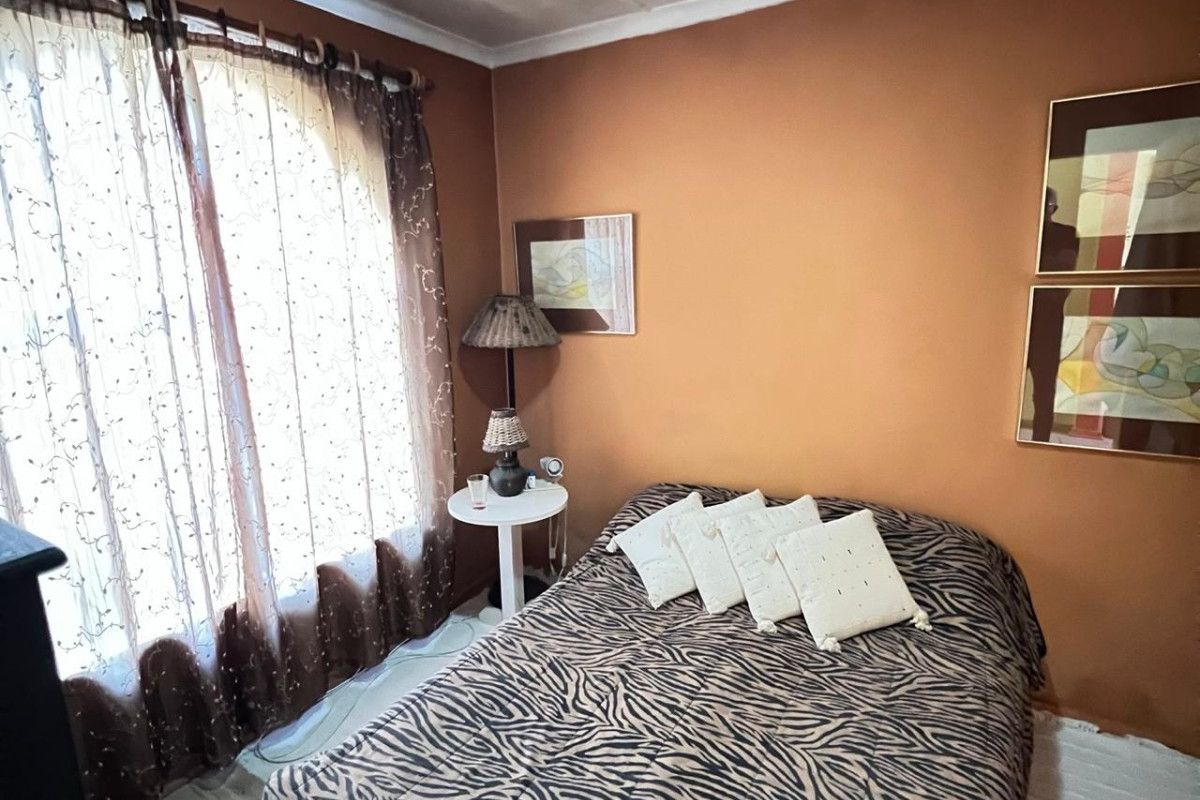 A bedroom featuring terracotta-toned walls and a large window with sheer curtains. The ceiling shows visible signs of staining.