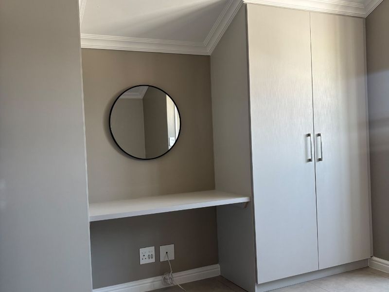 A clean, modern bedroom featuring built-in cupboards, a dedicated desk area, crown molding, and neutral tiled flooring.